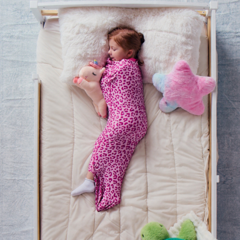 Child sleeping on a cream bed sheet while wearing a pink leopard print Junior Sleep Pod and holding a plush unicorn.